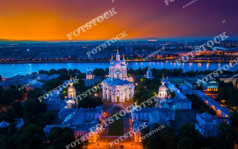Панорамный вид с дрона на Смольный собор в Санкт-Петербурге вечером, на фоне реки и огней ночного города. Яркая подсветка куполов и фасада собора, отражения в воде и городской ландшафт создают впечатляющий ночной пейзаж северной столицы России. Аэрофотосъёмка подчёркивает величие архитектуры, историческую значимость здания и атмосферу ночного Петербурга. Изображение идеально подходит для публикаций о путешествиях по России, архитектуре Санкт-Петербурга, аэрофотосъёмке, достопримечательностях города и городских панорамах.